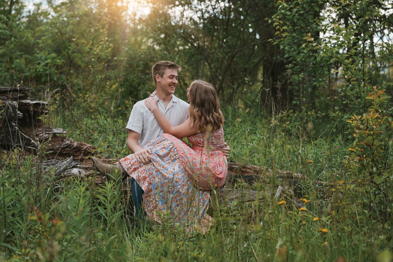 A romantic couple sits in a sunny meadow surrounded by tall grass and wildflowers during golden hour.