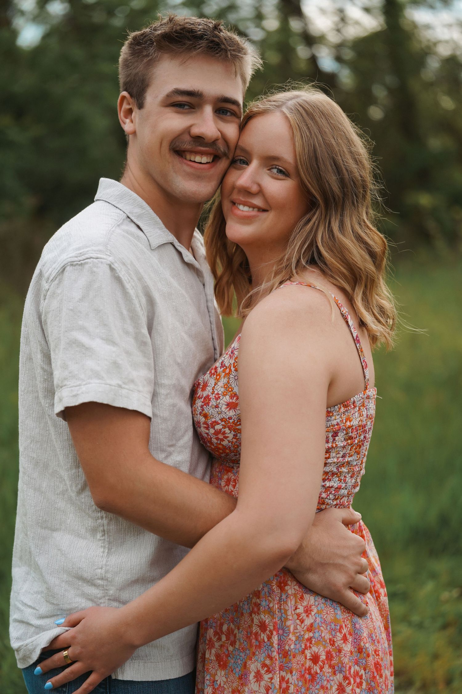 Couple embraces and smiles together outdoors in casual summer outfits with green foliage in background.