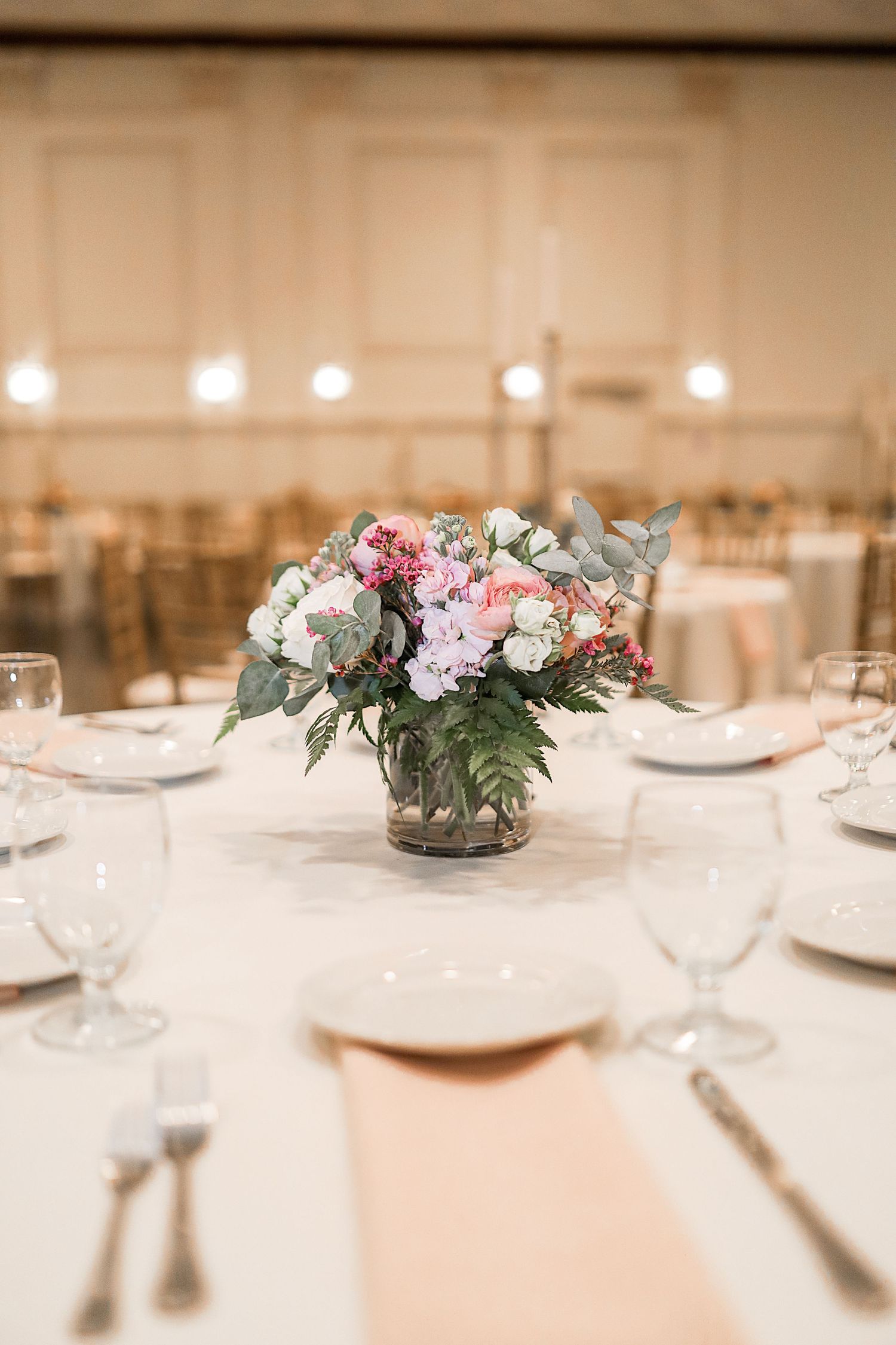 Elegant wedding reception table settings with pink and white floral centerpieces and crystal glassware in soft lighting.