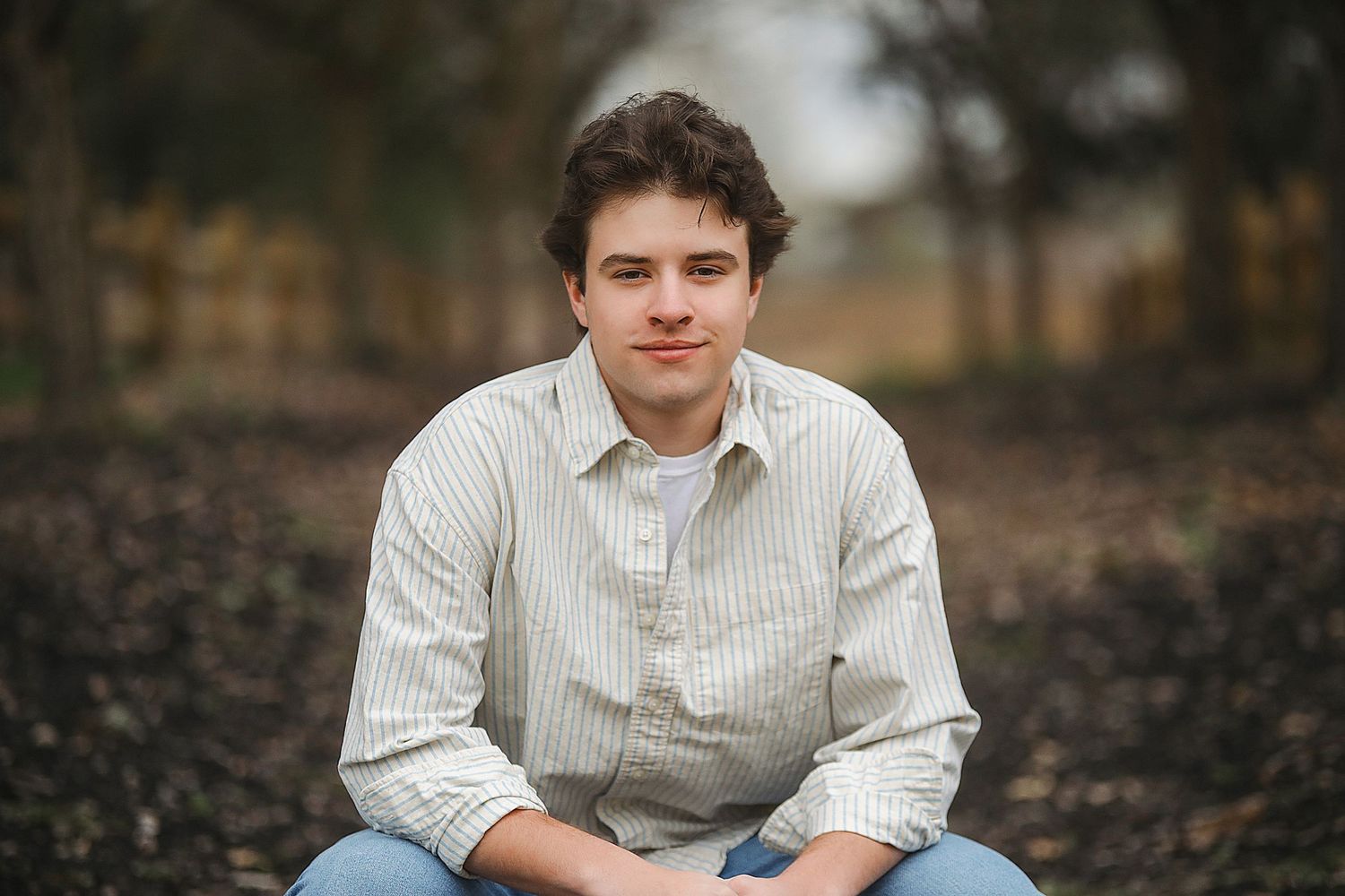 A person in a striped button-down shirt sits casually outdoors on a blurred natural background.