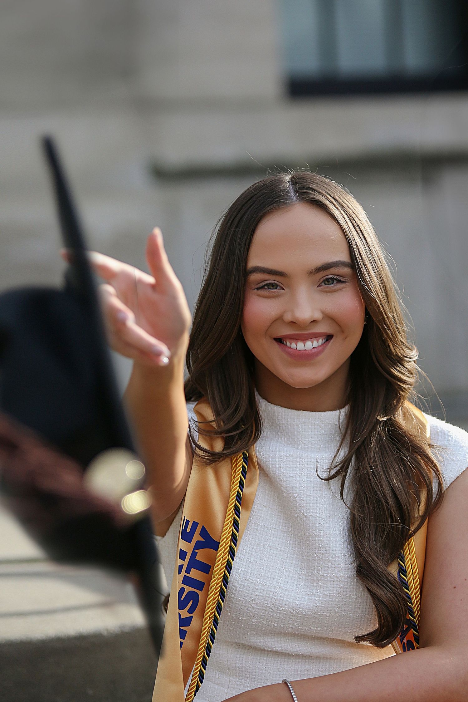 A person in a white sleeveless top smiles and waves while holding a yellow and blue graduation stole.