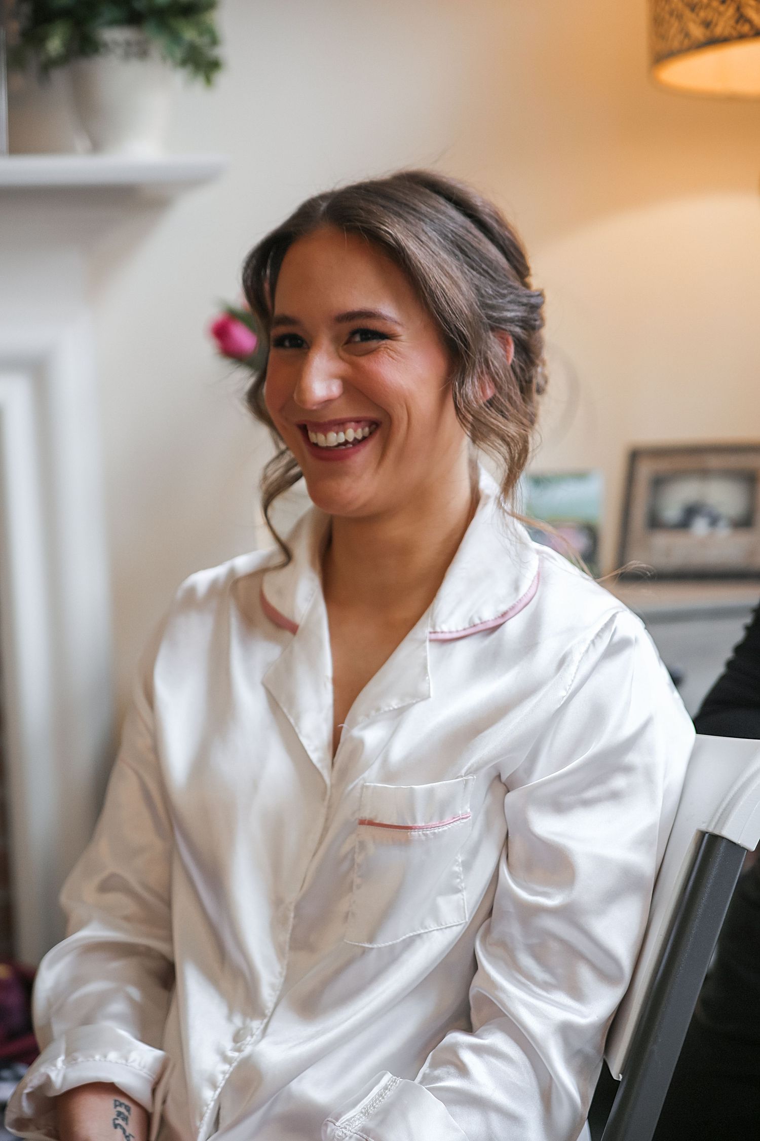A person wearing a white satin robe sits in a chair while smiling and laughing in a warmly lit room.