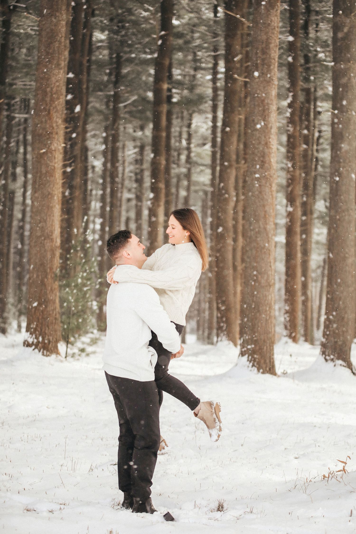 A couple playfully throws snow at each other in a snowy winter forest during an engagement photoshoot.