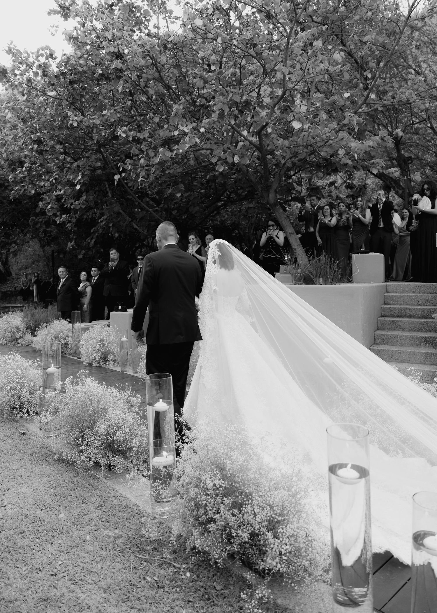 Black and white photo of bride in long veil walking down outdoor aisle with escort at elegant garden wedding ceremony.