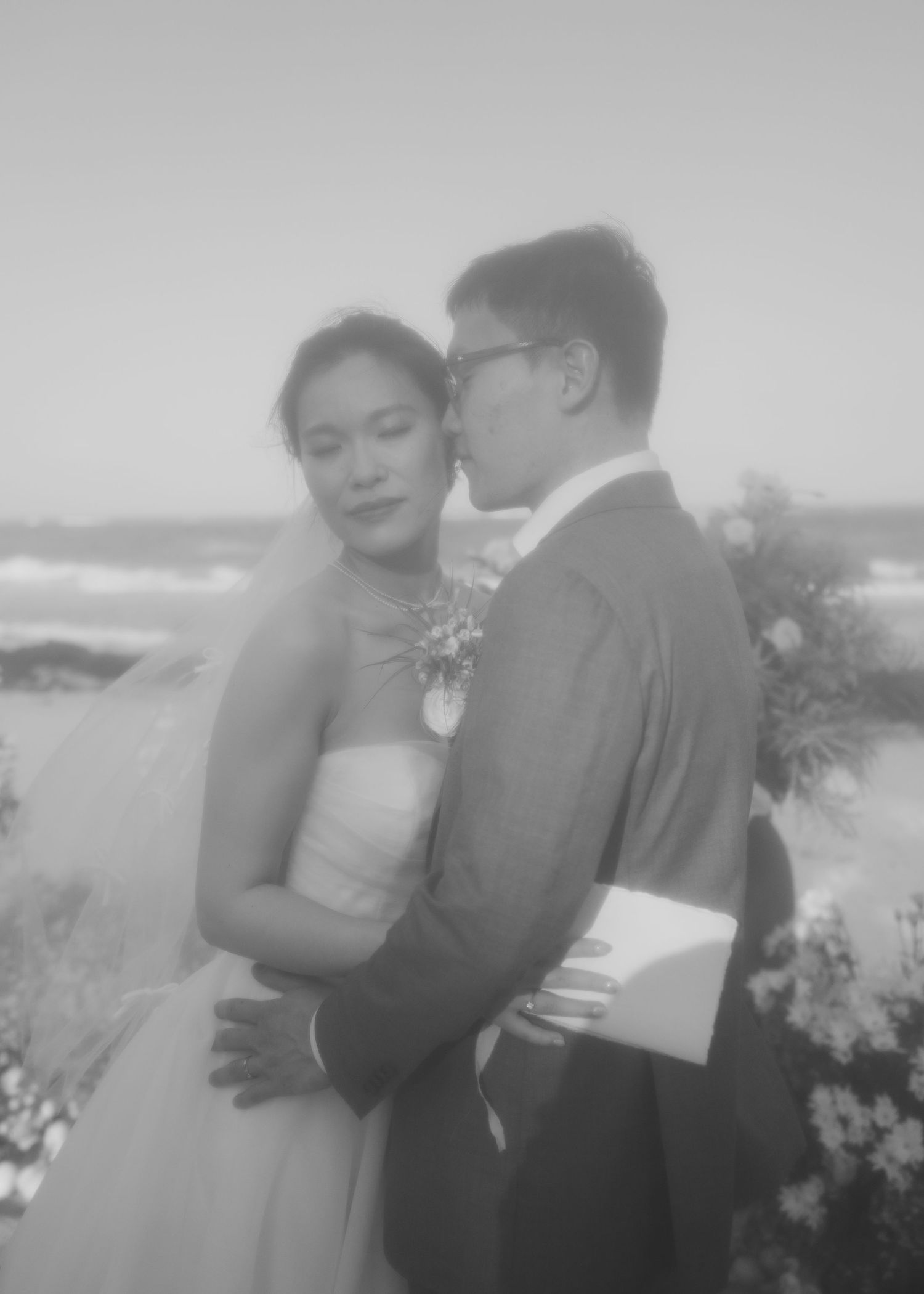 Black and white photo of a couple embracing at a beach wedding, groom in suit kissing bride in strapless gown.