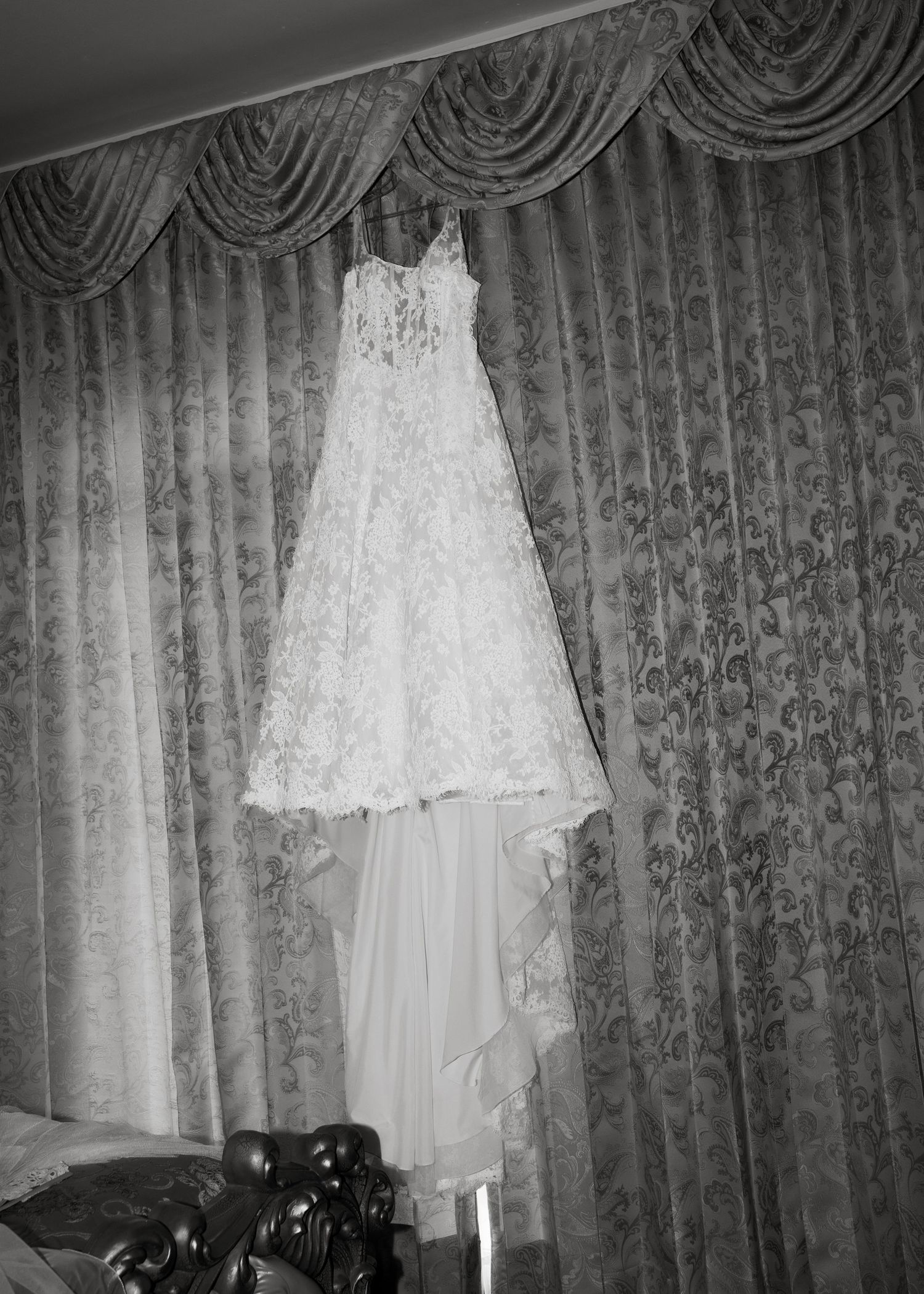 A white lace wedding dress hangs elegantly against ornate curtains in a monochrome photograph.