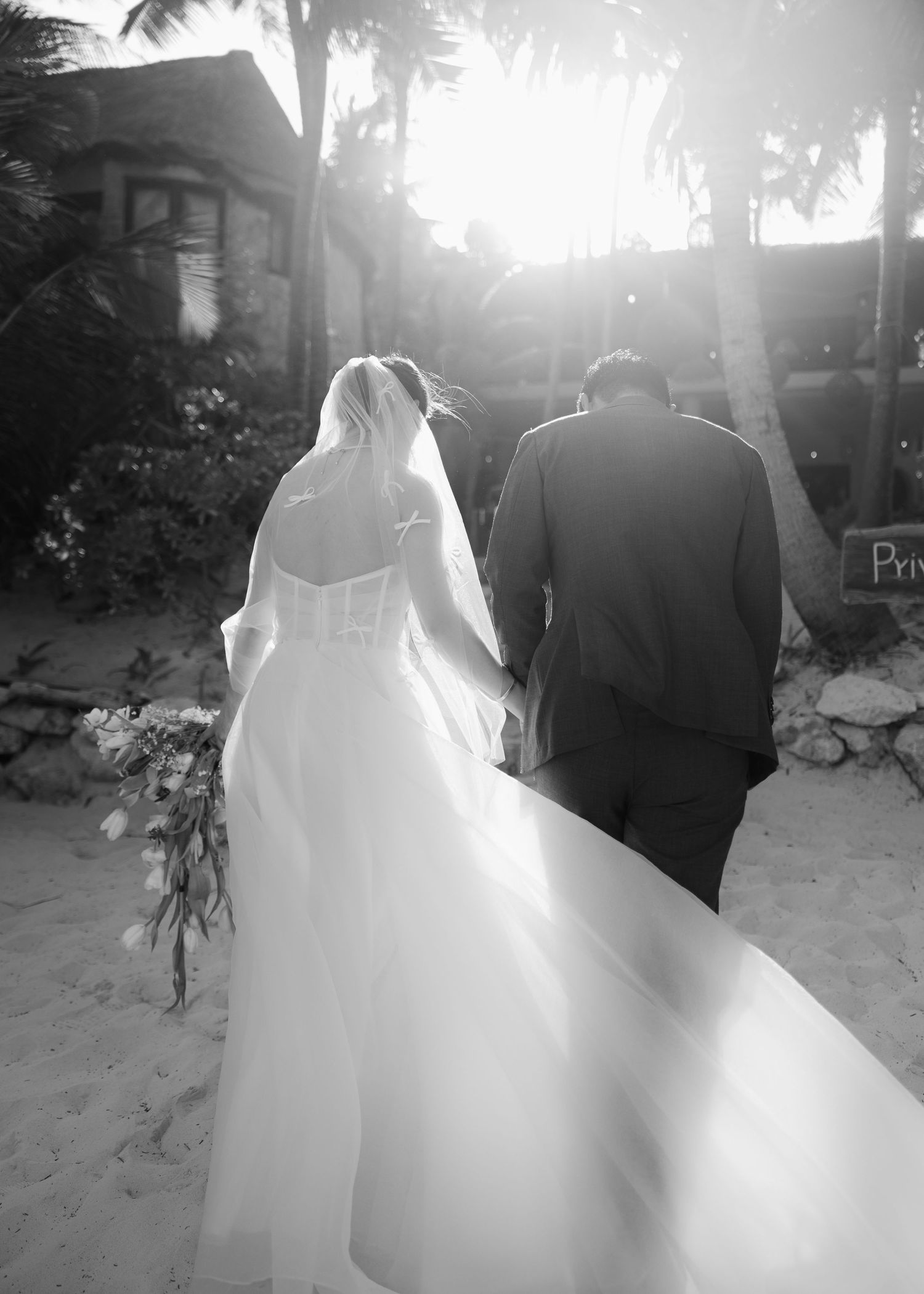 Black and white photo of a bride and groom walking hand in hand on a beach, with palm trees and sunlight in the background.