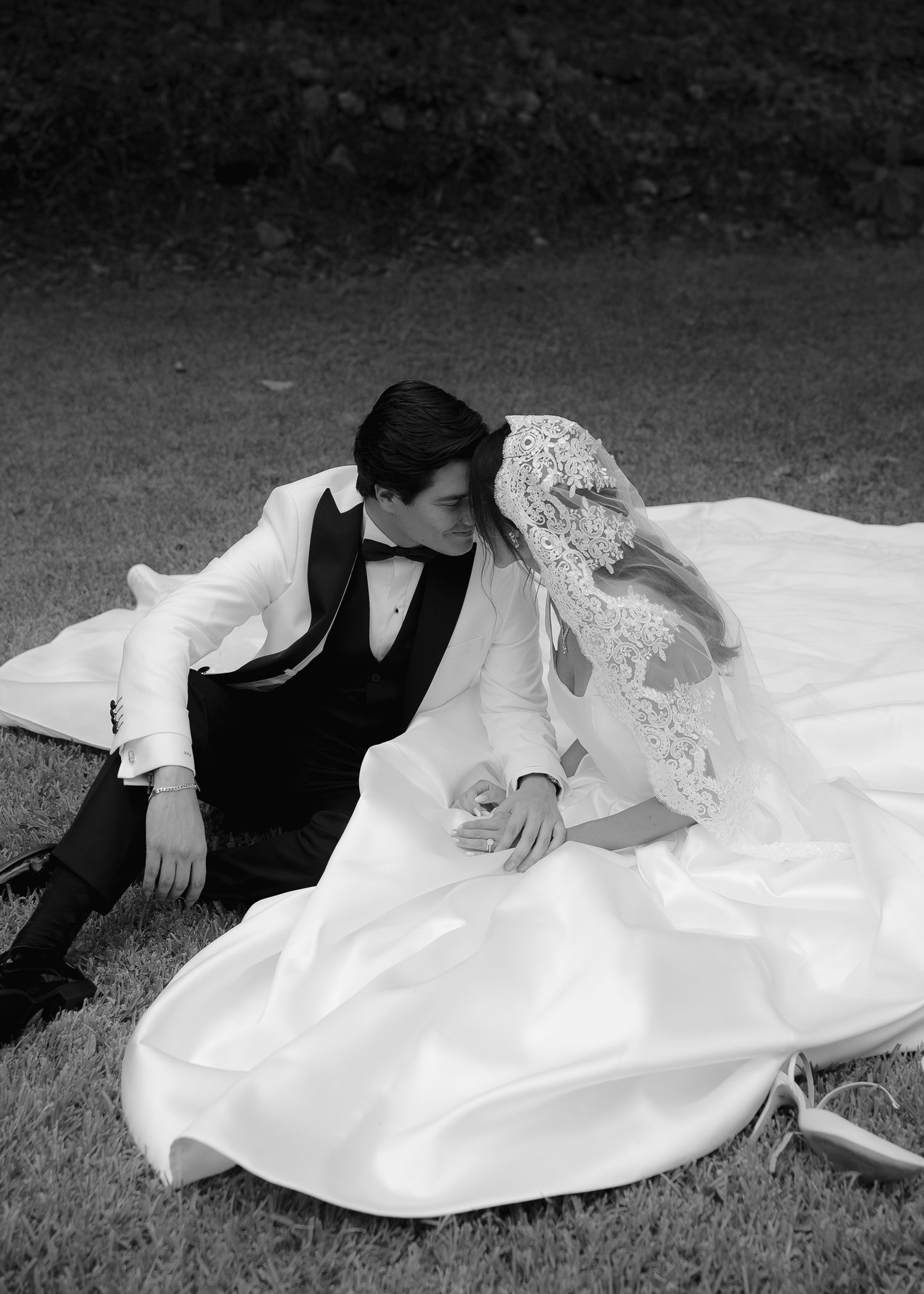 Black and white photo of a bride and groom sitting on grass, sharing a romantic moment in wedding attire.