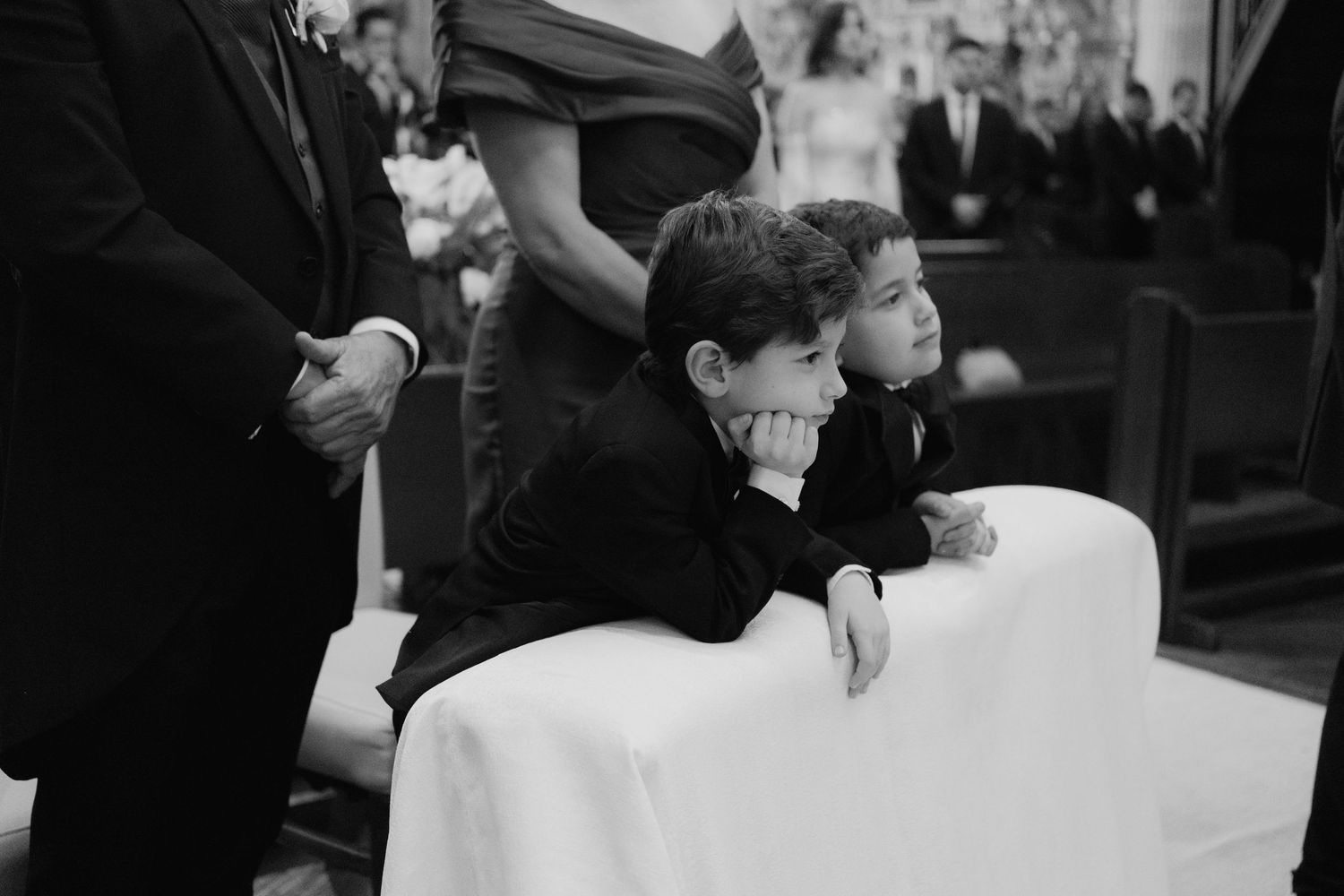 Two young boys in black suits sit bored at a wedding ceremony, leaning on a white pew in a black and white photo.
