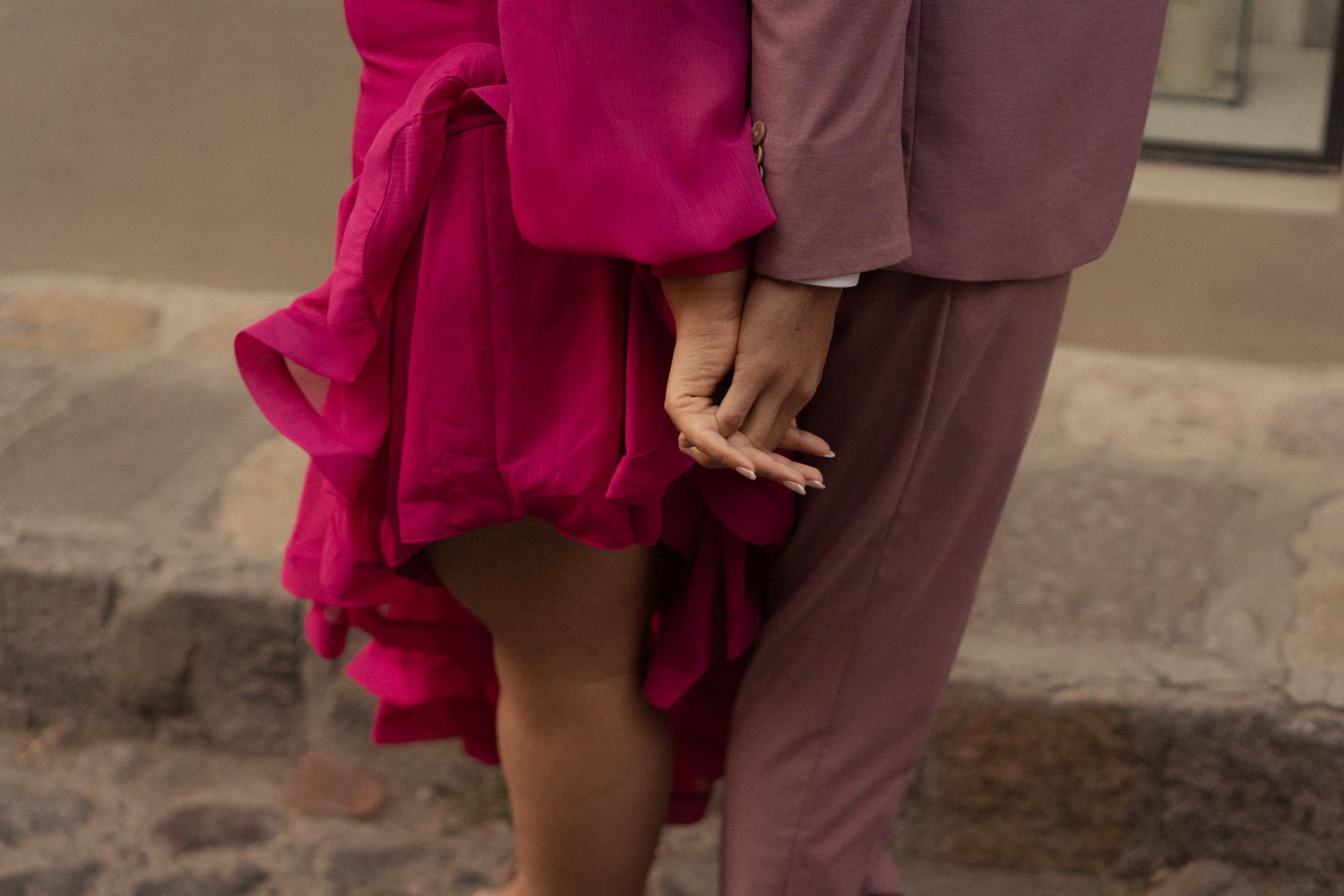 Two people holding hands, one wearing a flowing fuschia dress and the other in brown pants, standing together.