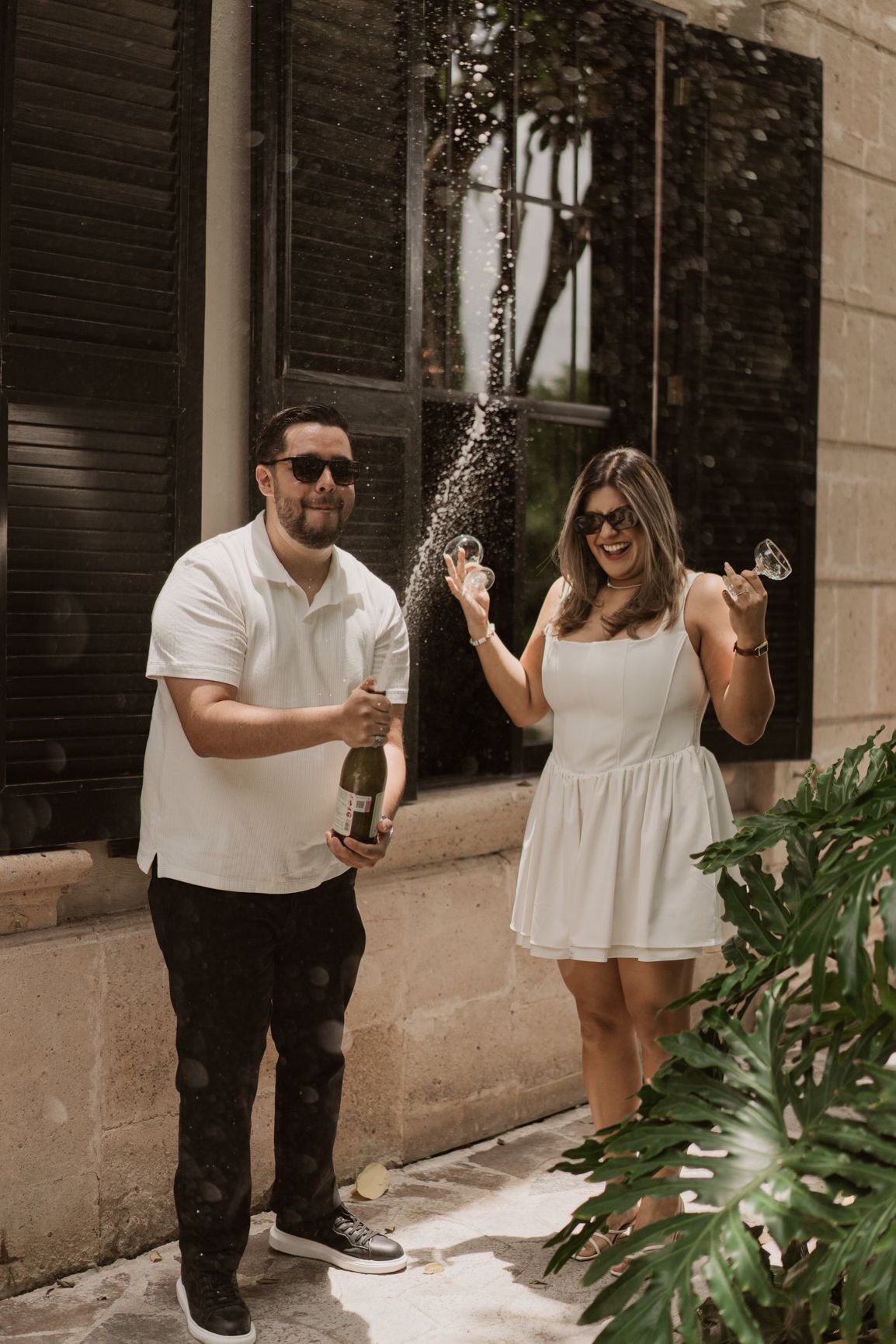 Romantic couple spraying champagne and celebrating together outdoors while dressed in black and white outfits.