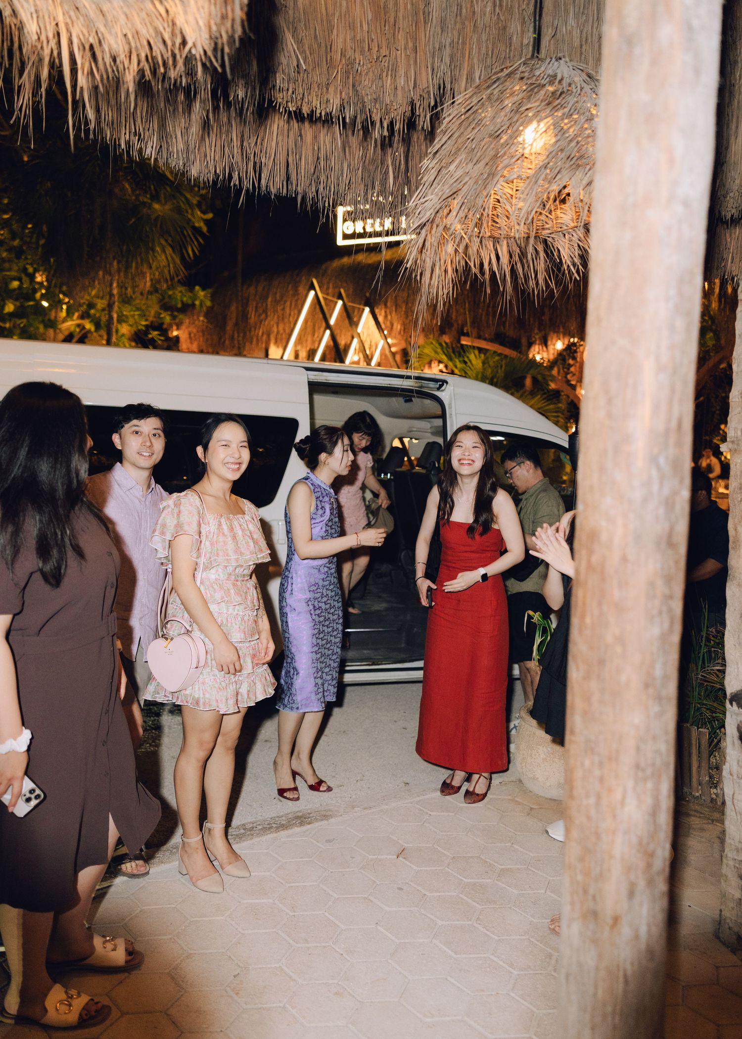 Guests in elegant attire gather outside a tropical venue at night, with a white van and lush palm decor.