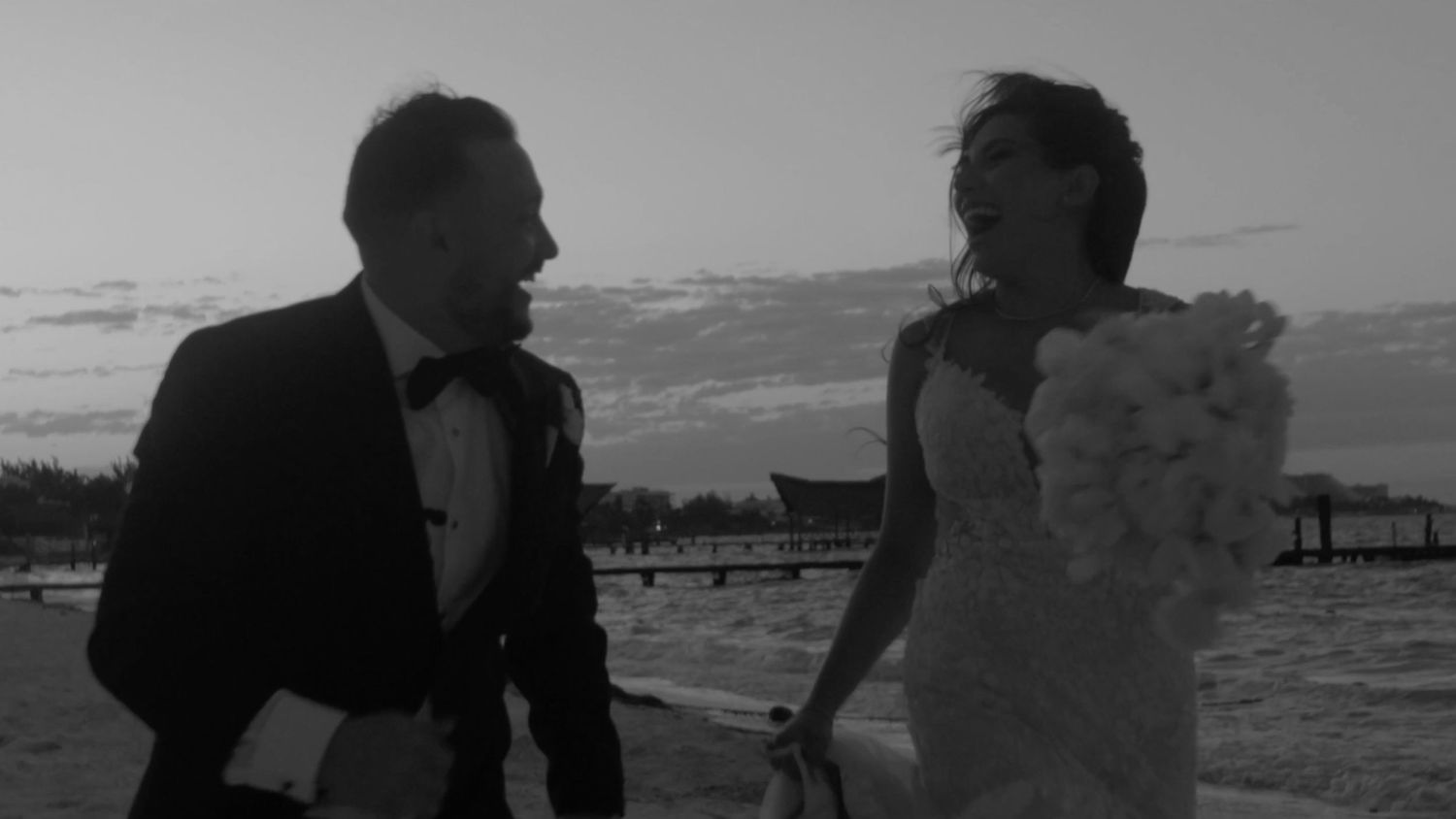 A black and white photo of a couple running along a beach at sunset in wedding attire.