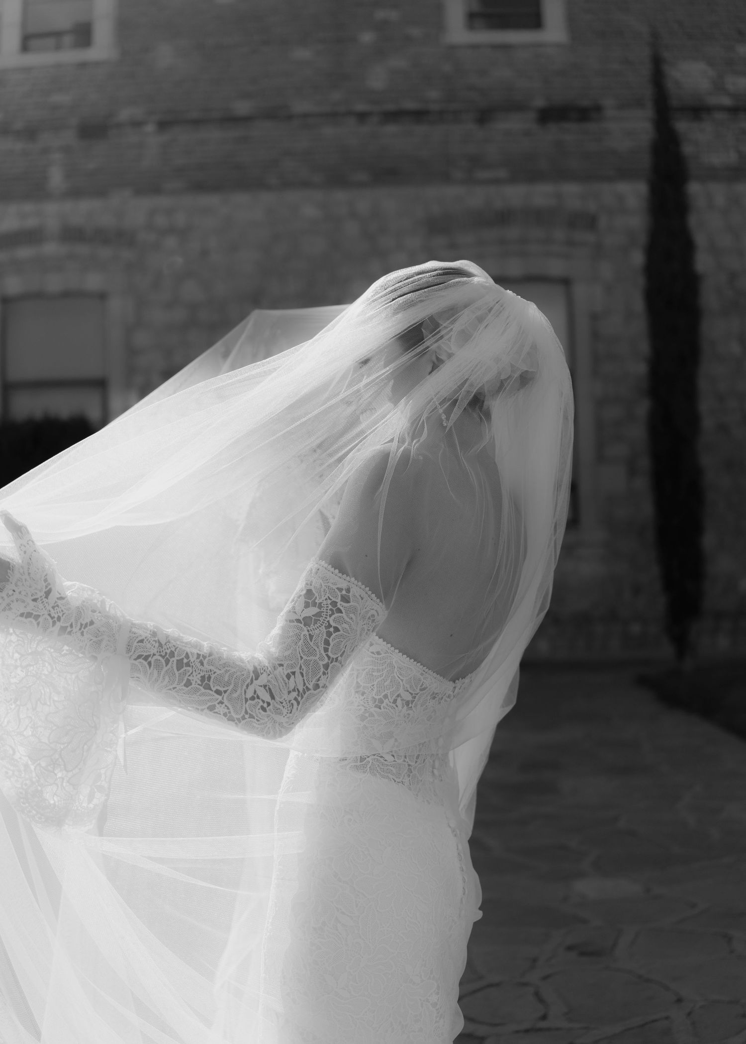 Black and white photo of a bride in lace wedding dress with veil blowing over her face outdoors near stone building.