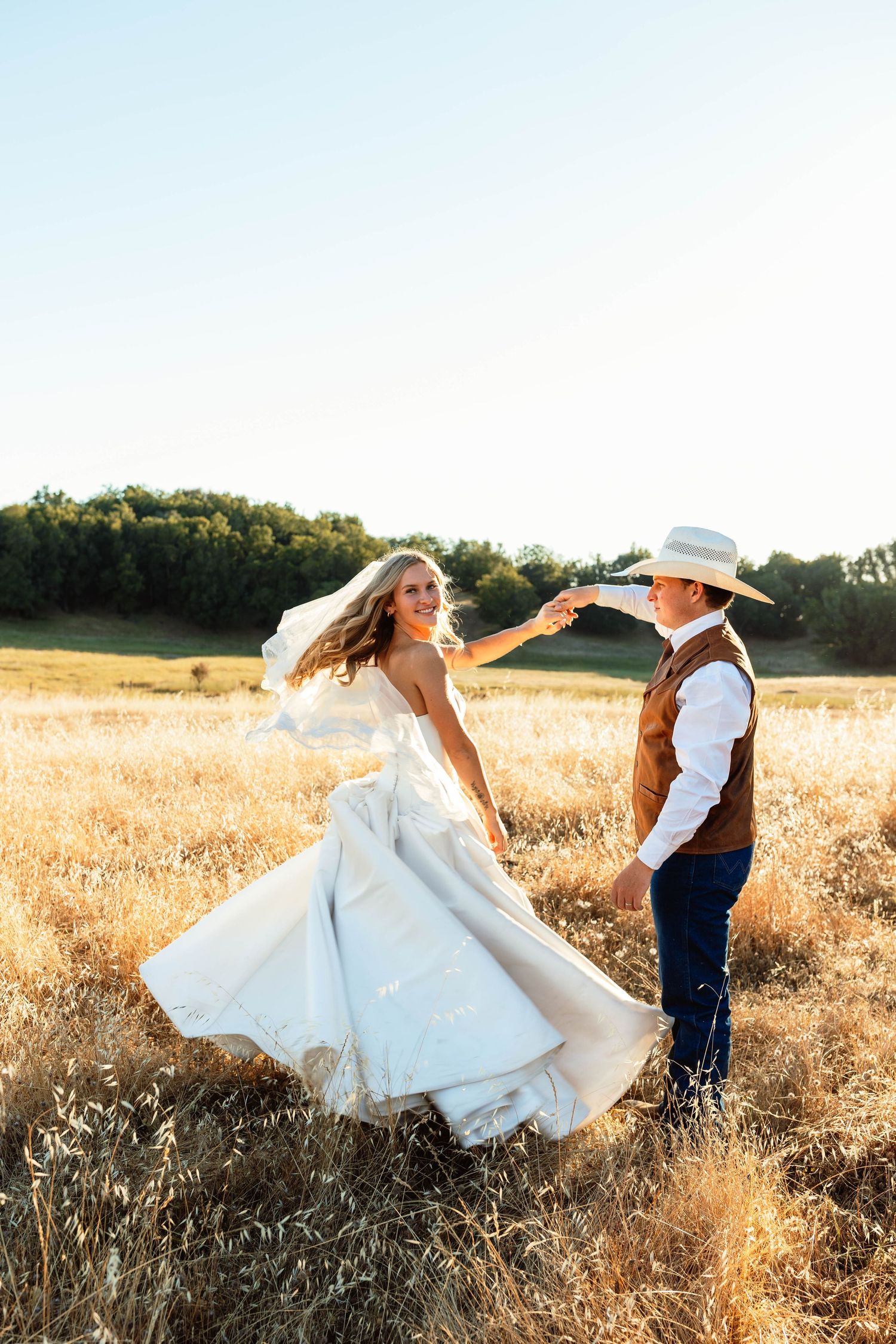Bride in white gown twirls with groom in cowboy hat in golden field at sunset.