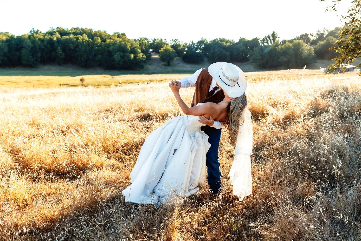 Couple sharing a romantic kiss in a golden field, bride in white dress and groom in cowboy hat, rustic outdoor wedding.