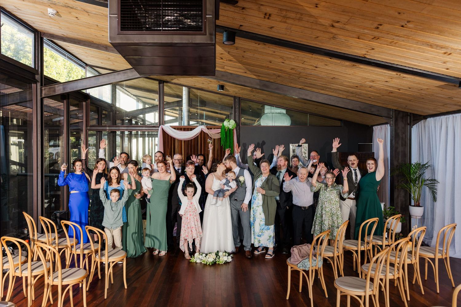 Wedding group photo with guests in green attire celebrating indoors at a modern venue with wood ceiling.