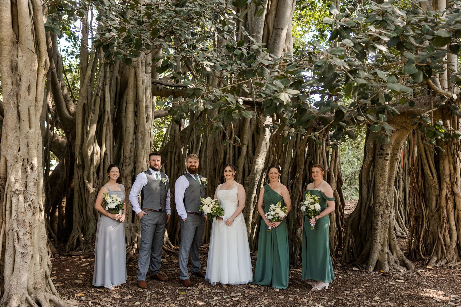 Wedding party poses before a massive banyan tree, bridesmaids in sage green and gray gowns flanking bride and grooms.