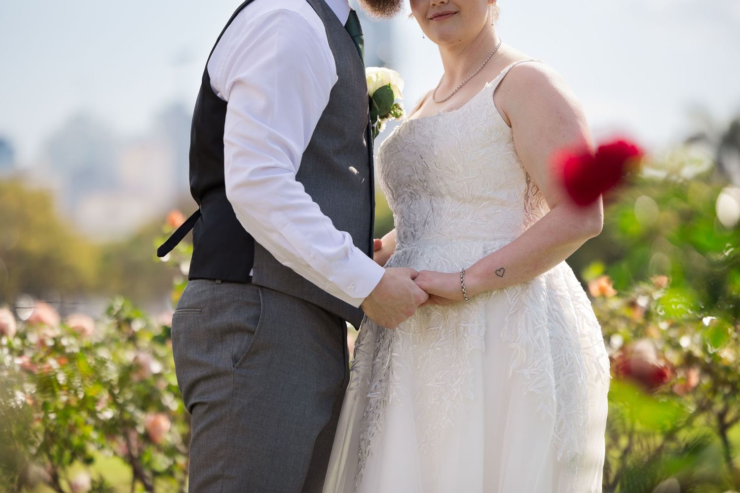 Bride and groom holding hands in a rose garden, bride wearing lace wedding dress holding red flowers.