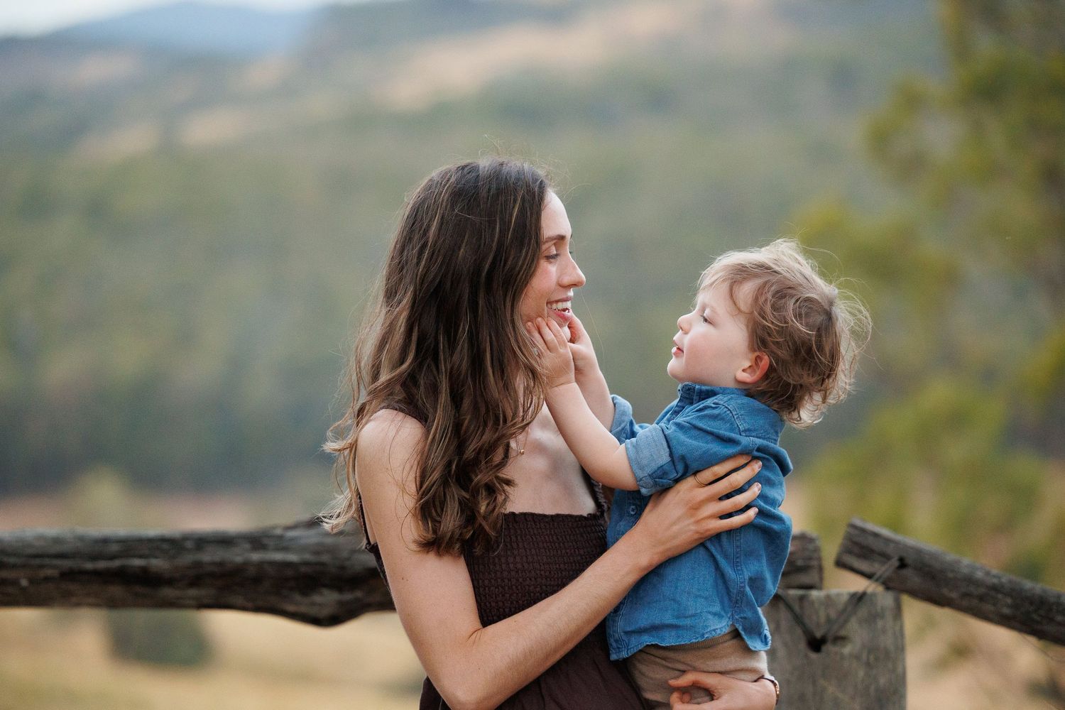 A young mother holds her toddler outdoors near a rustic wooden fence with green mountain views.