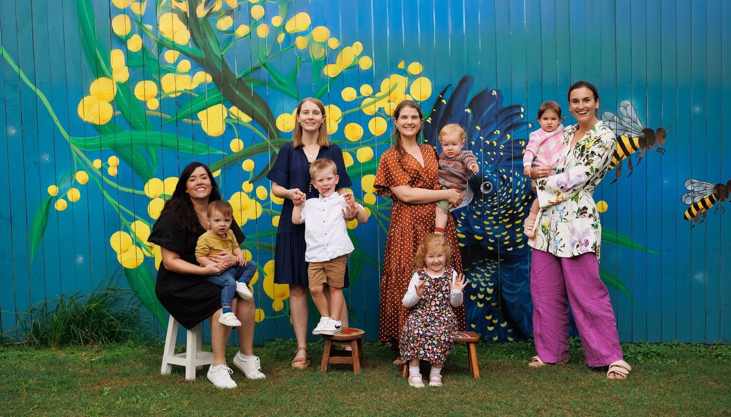 Group of mothers with children posing in front of colorful floral mural with bees and wattle flowers.