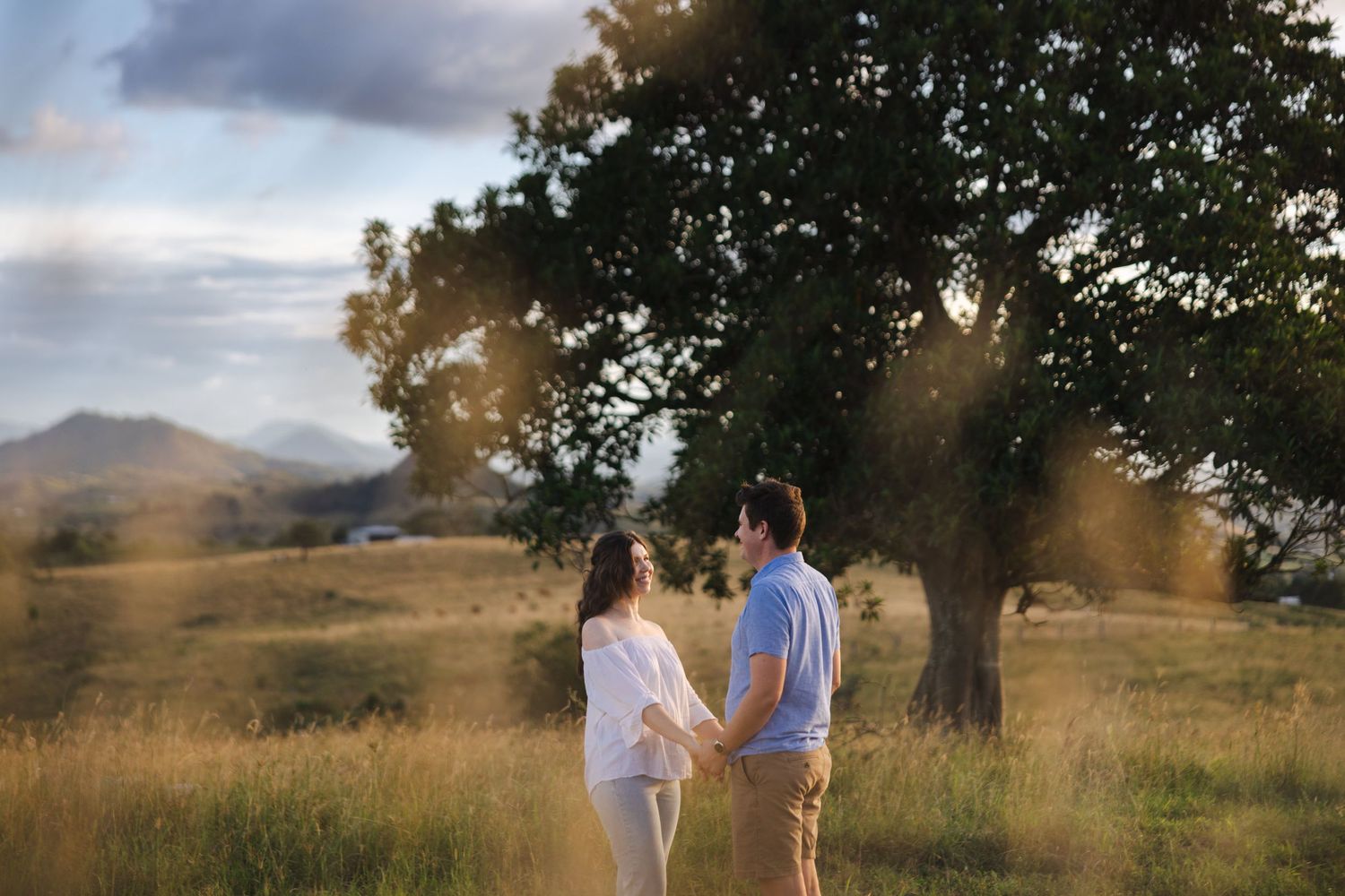 Two people stand together in a golden field at sunset with mountains and trees silhouetted in the background.