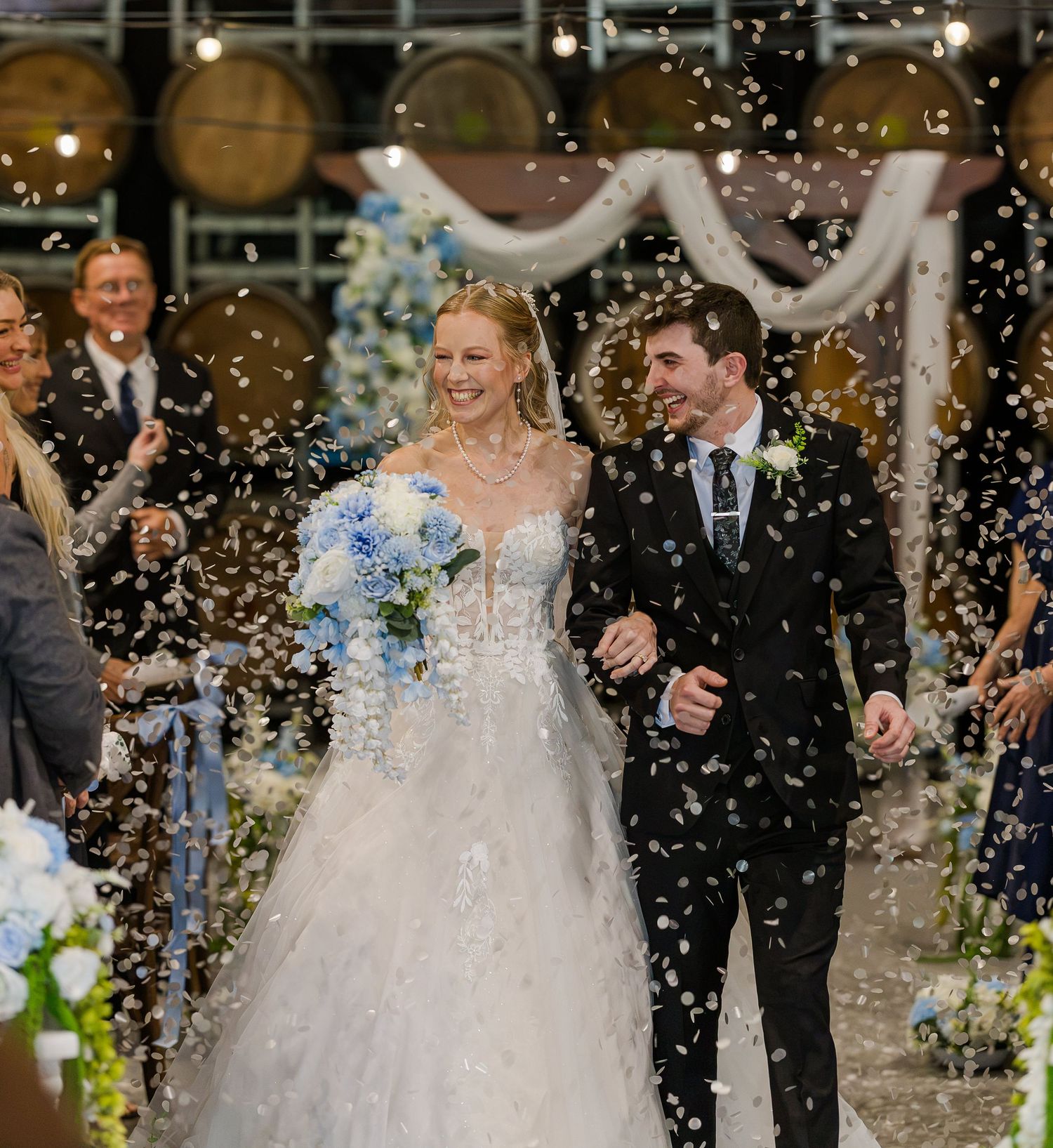 Bride and groom walk down the aisle as guests shower them with confetti at elegant indoor wedding ceremony.