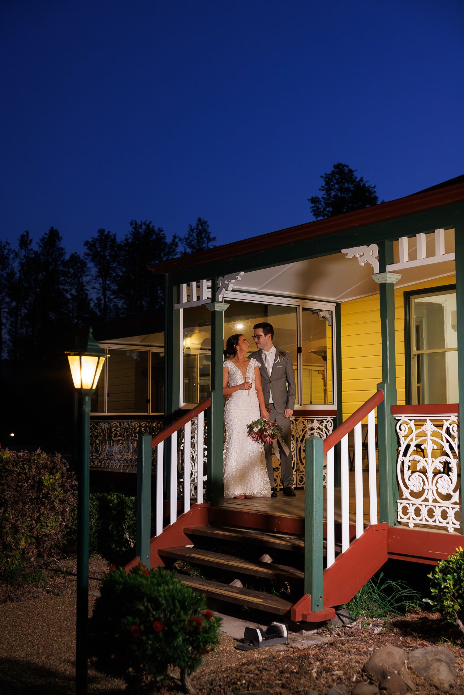 Bride and groom share a romantic moment on a colorfully lit Victorian porch at dusk.
