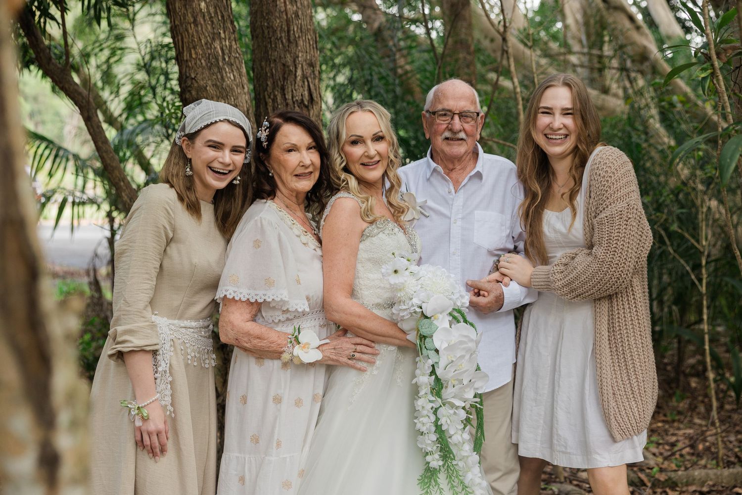 A bride in a white dress holds a floral bouquet surrounded by smiling family members in a lush outdoor setting.