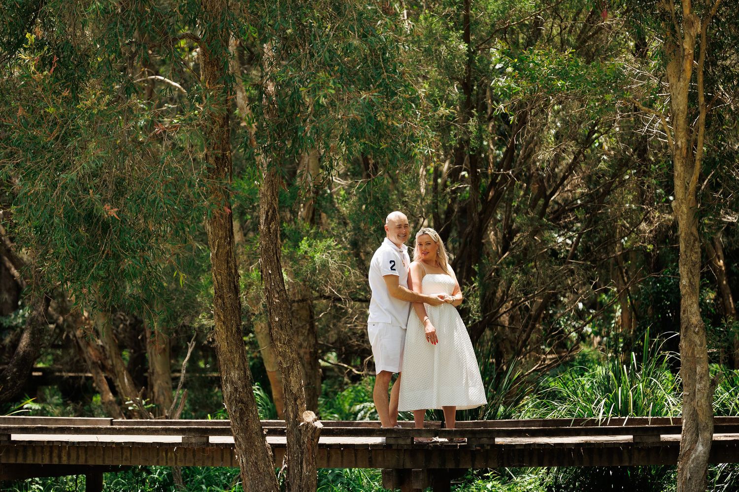 Two people in white dresses stand together on a wooden walkway surrounded by lush green trees in a garden setting.