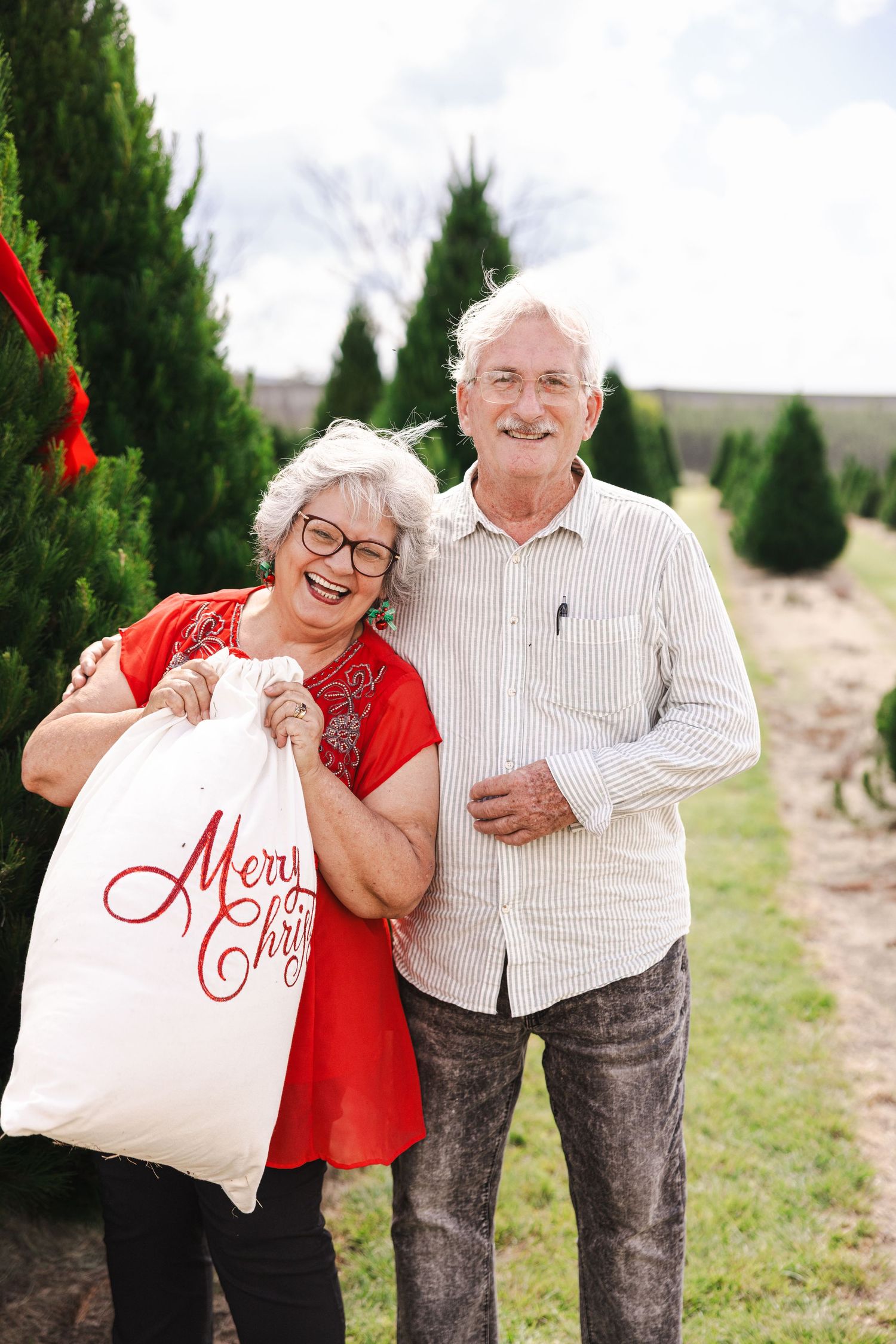 Elderly couple shares joyful moments at a Christmas tree farm while holding a festive tote bag.