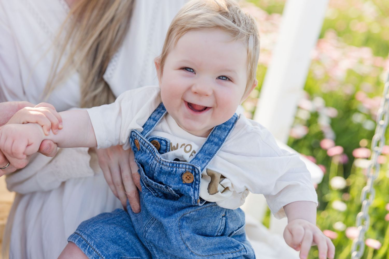 Smiling baby in denim overalls and white shirt held by mother outdoors among pink flowers.