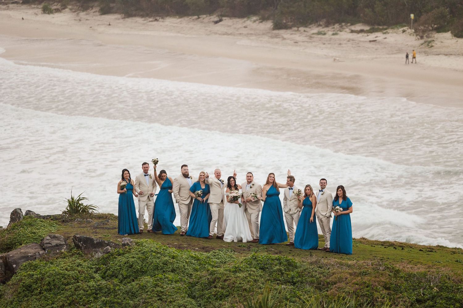 Wedding party in teal gowns and tan suits posing on a coastal cliff with ocean waves and sandy beach in the background.