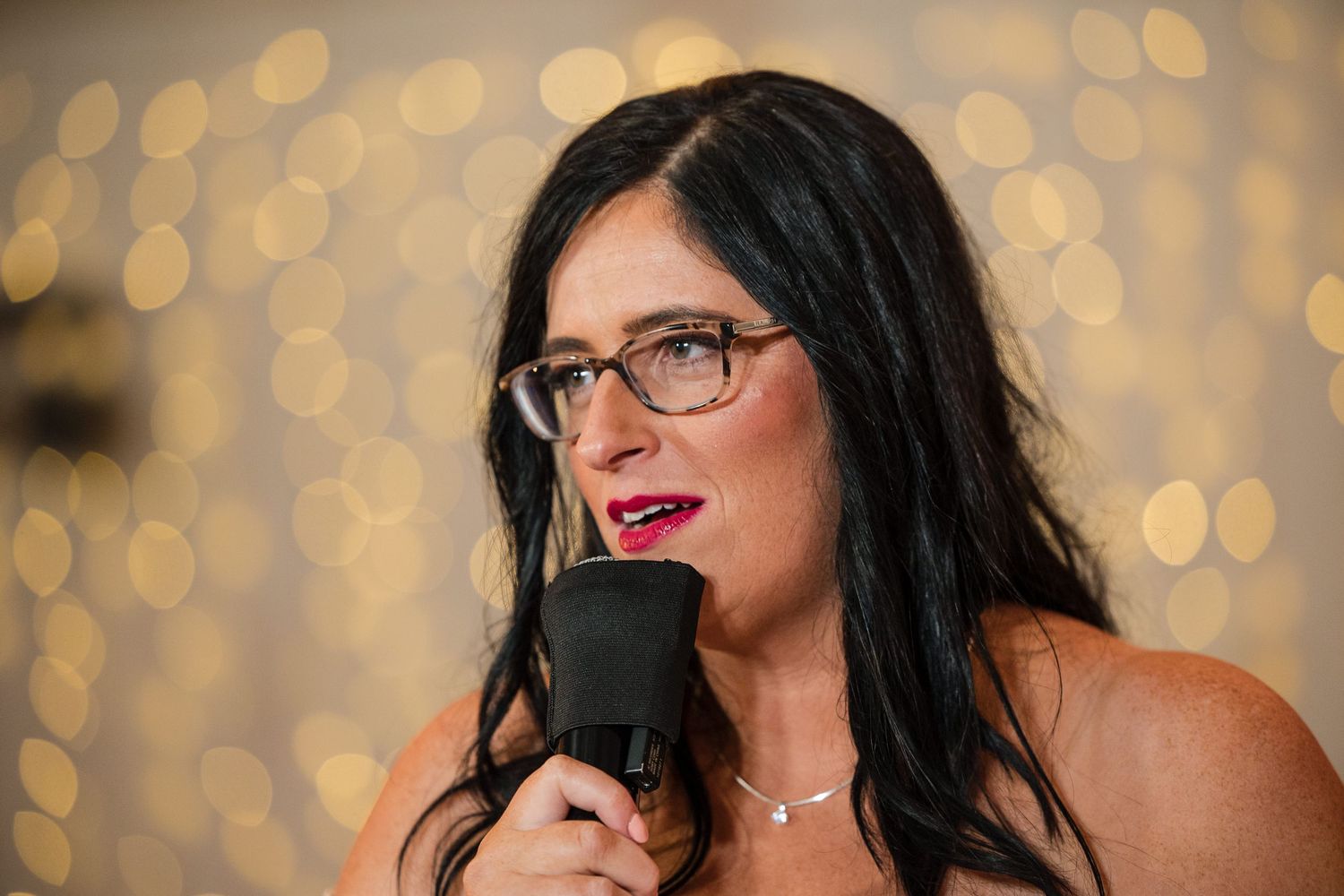Woman with dark hair and glasses speaking into a microphone at an elegant event with bokeh lights background.