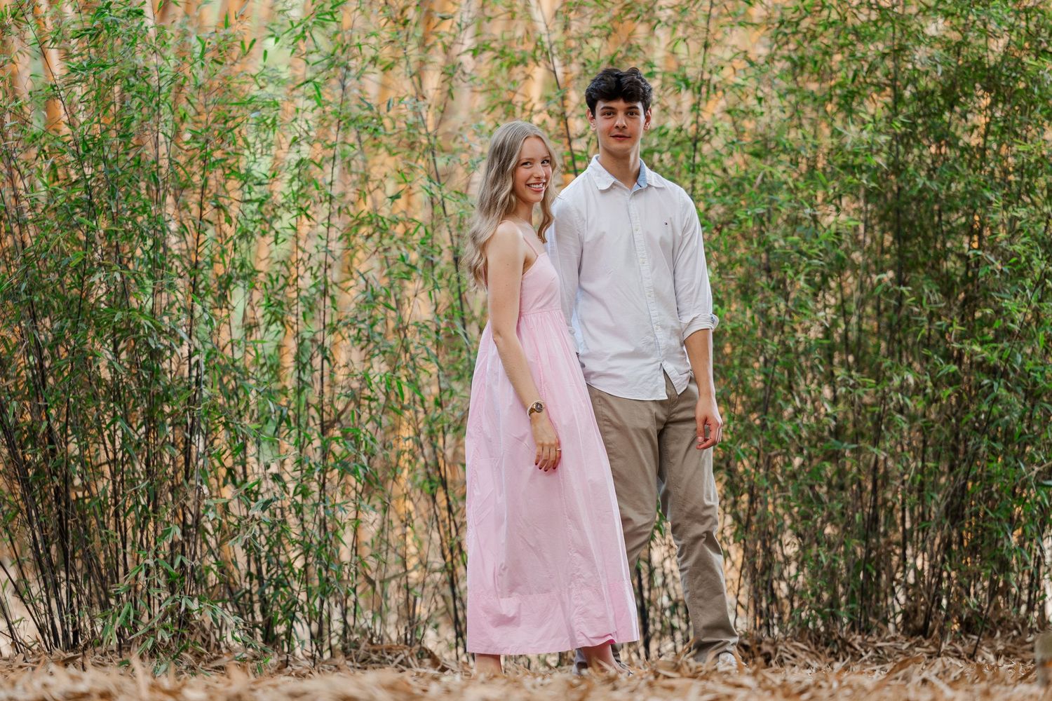A couple stands together in front of a bamboo grove, wearing a pink maxi dress and white button-down shirt.