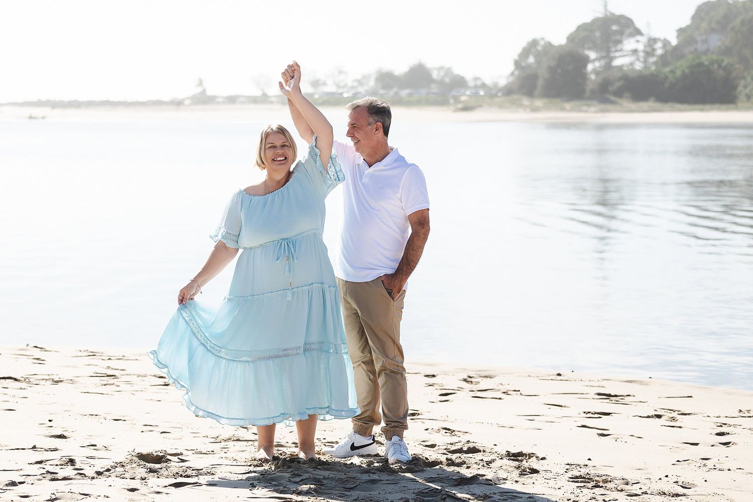 A couple dancing together on a serene beach with calm waters and trees in the background.
