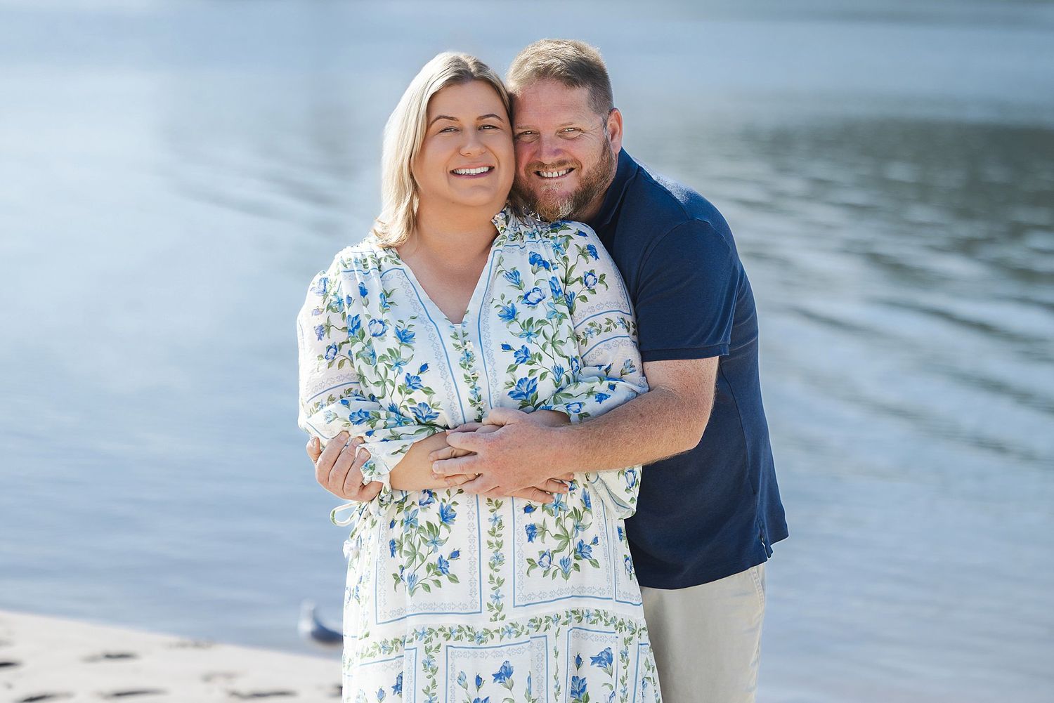 A couple poses in loving embrace on a sunny beach with the ocean waves in the background.