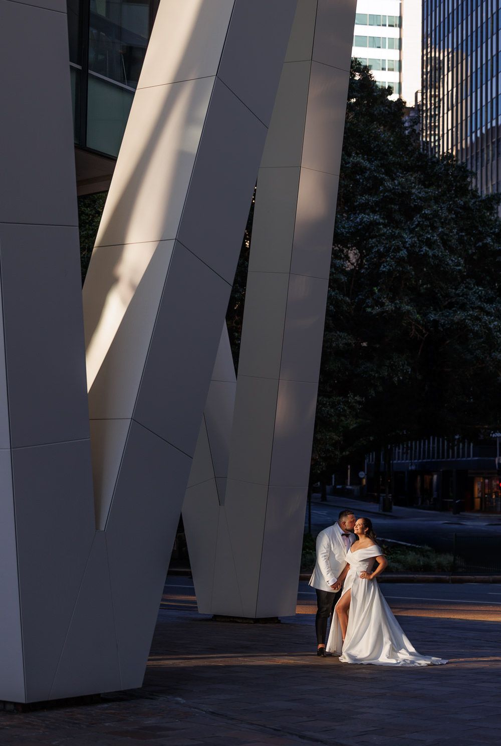 A couple embraces against a large white architectural sculpture in an urban setting with dramatic lighting and shadows.