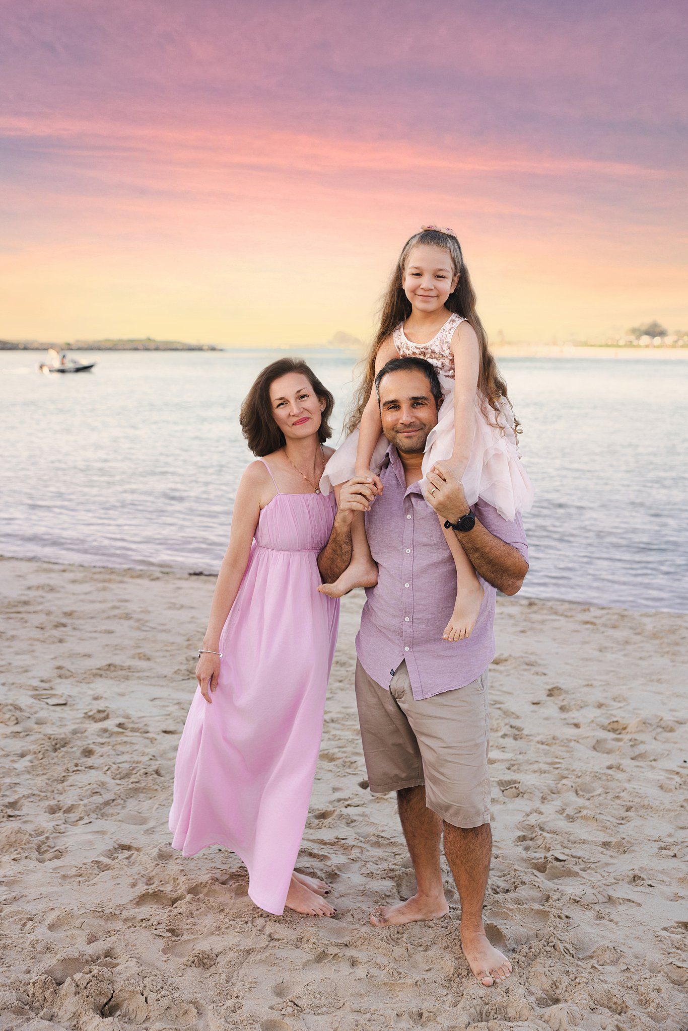 A family poses together on a beach at sunset wearing coordinating pink and beige outfits.