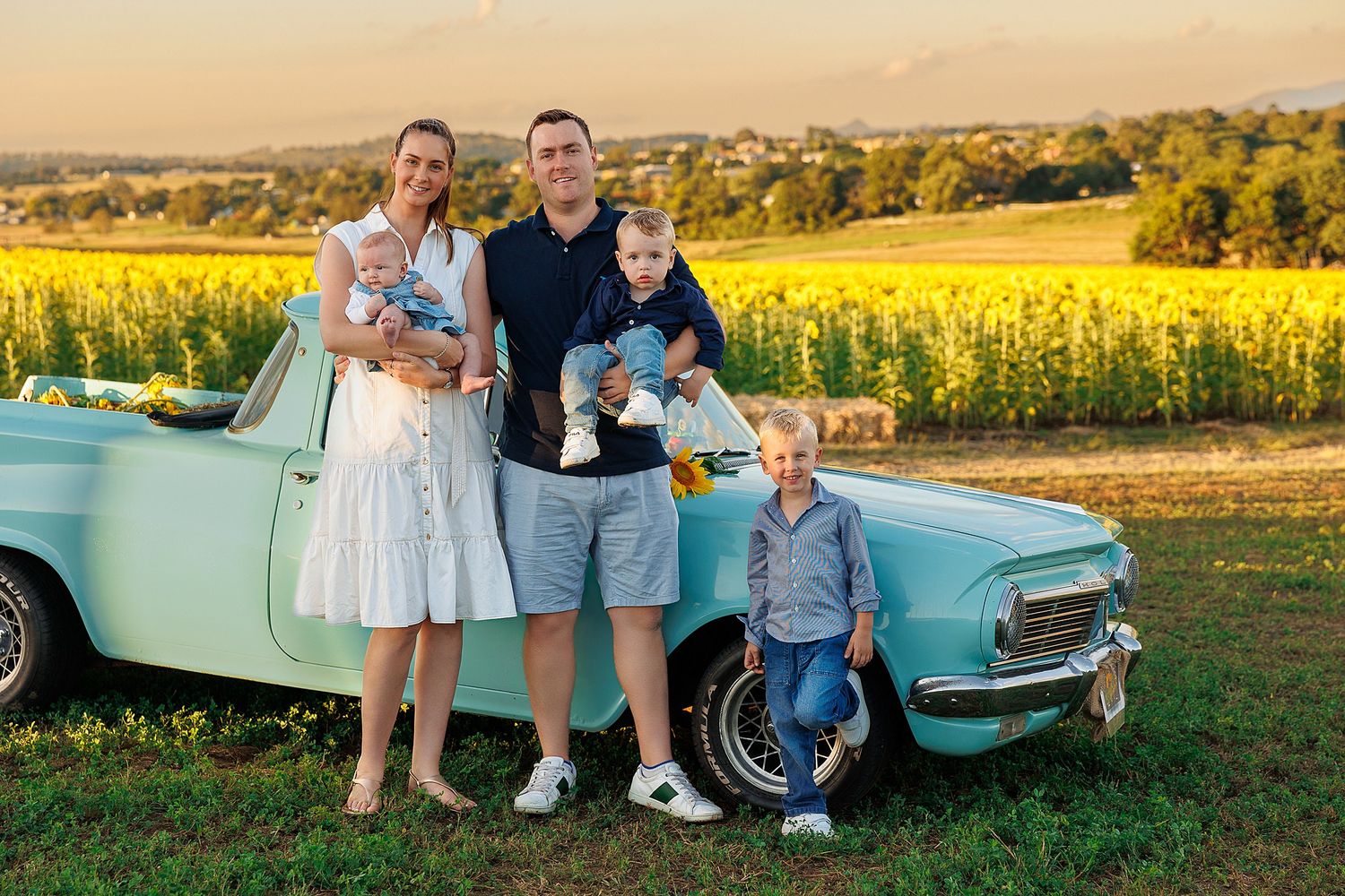 A family poses by a vintage mint green car in a field of yellow flowers at sunset.