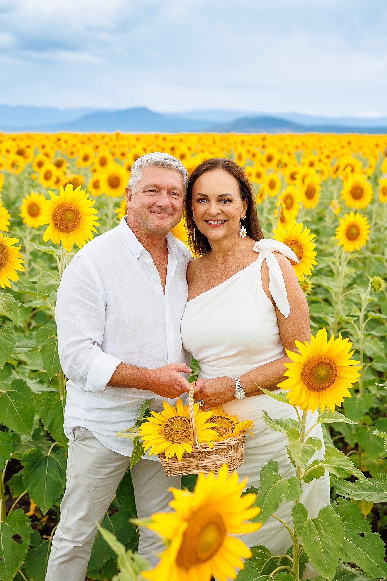 Couple in white outfits posing together in a vibrant sunflower field with mountains in the distant background.