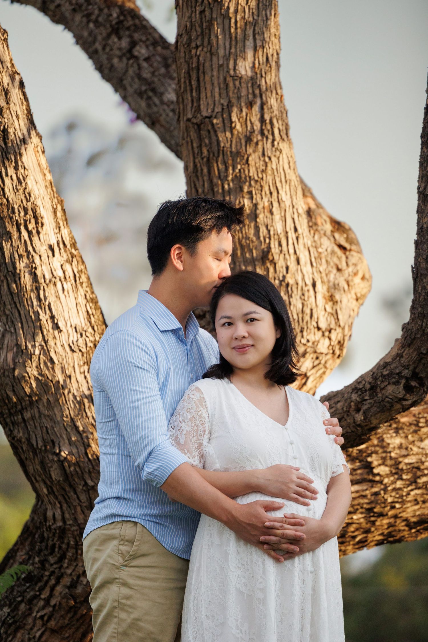 A couple embraces under a large tree trunk, with the person in white wearing a lace dress during a maternity photoshoot.