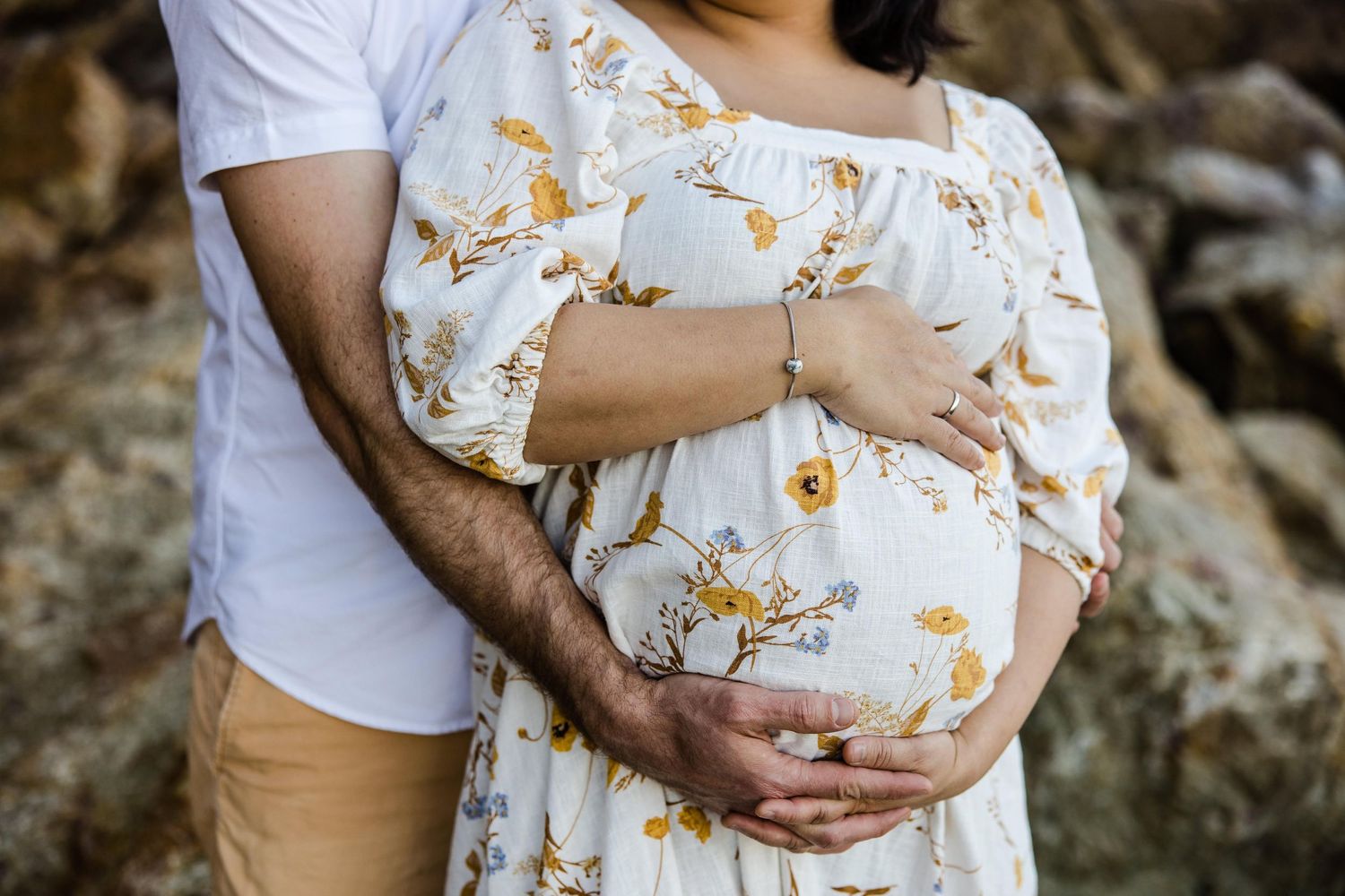 Close-up of couple embracing during maternity photo shoot wearing white and floral dress against rustic backdrop.