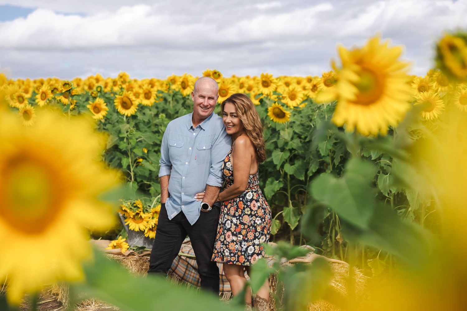 Couple smiling together in a vibrant sunflower field under a cloudy sky.