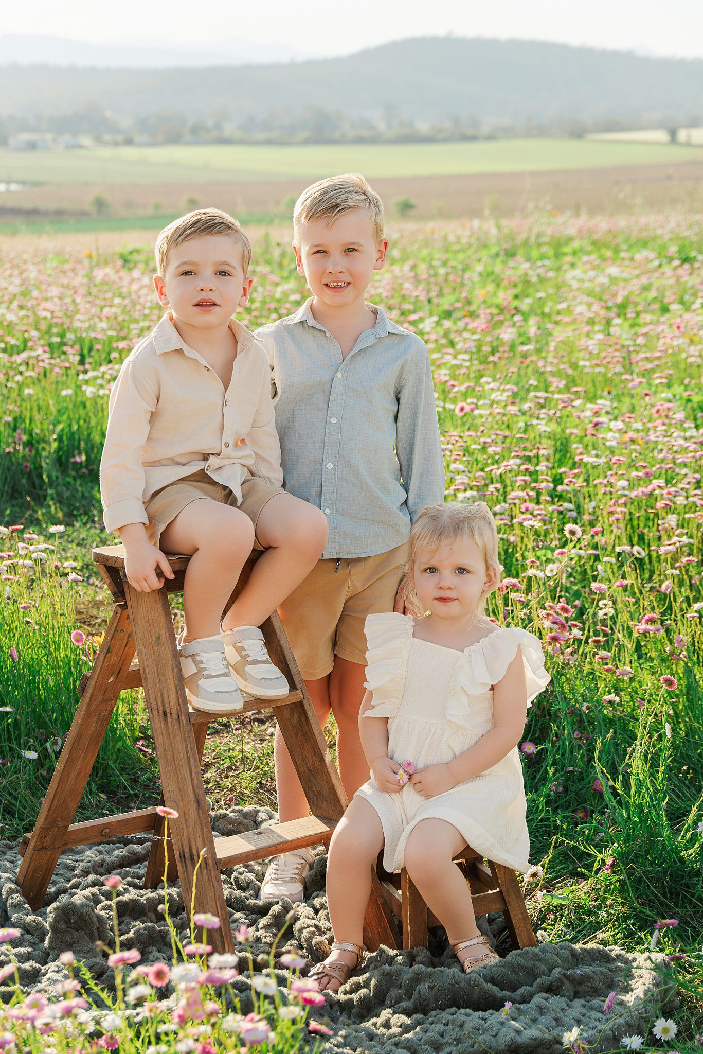 Two boys and a toddler girl in matching neutral outfits pose on wooden stools in a blooming wildflower field with rolling hills behind.