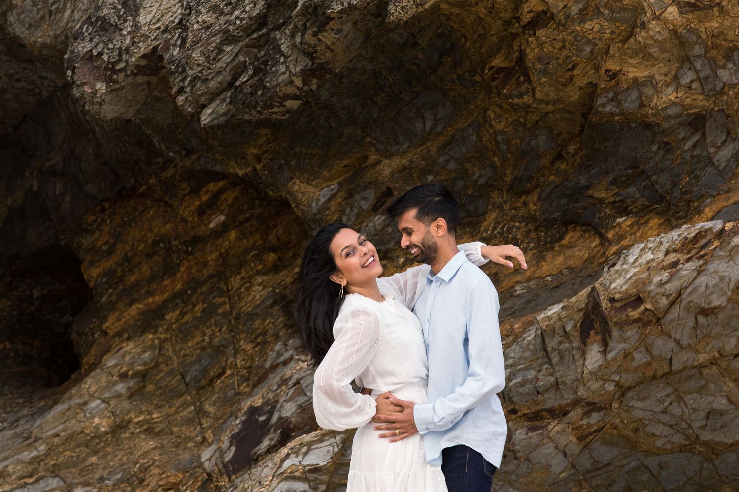 A couple in white shirts embraces while standing against rugged coastal rocks at sunset.