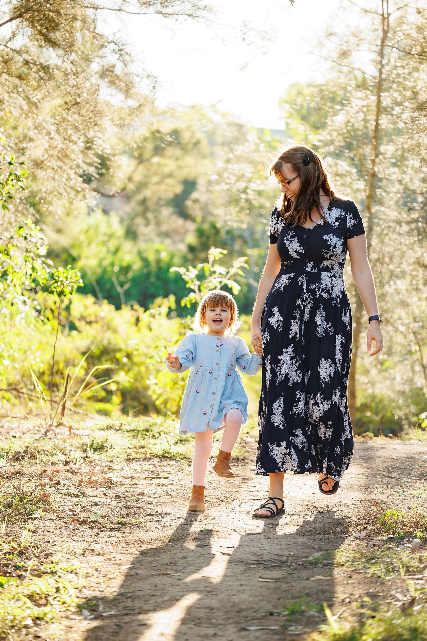 A pair walks together along a sunlit forest trail on a bright summer day, one wearing a floral maxi dress and the other in blue.