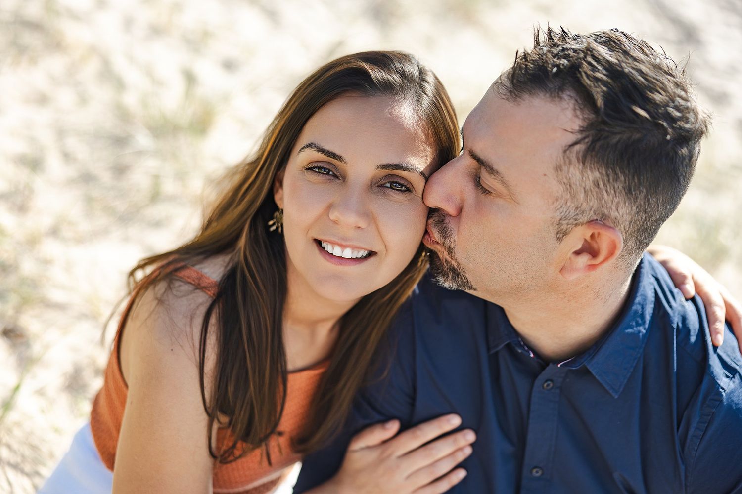 Romantic couple embracing outdoors in warm sunlight with woman in orange top and man in navy blue shirt.