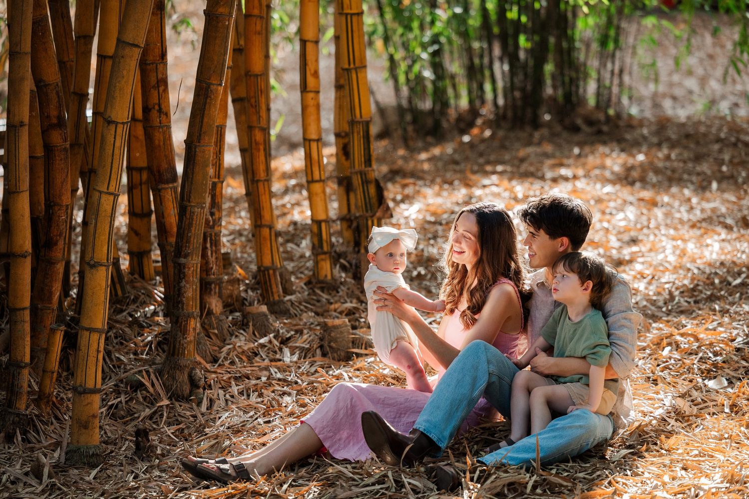 Two people sitting together on ground near bamboo trees while looking at mobile phones in a sunlit forest setting.