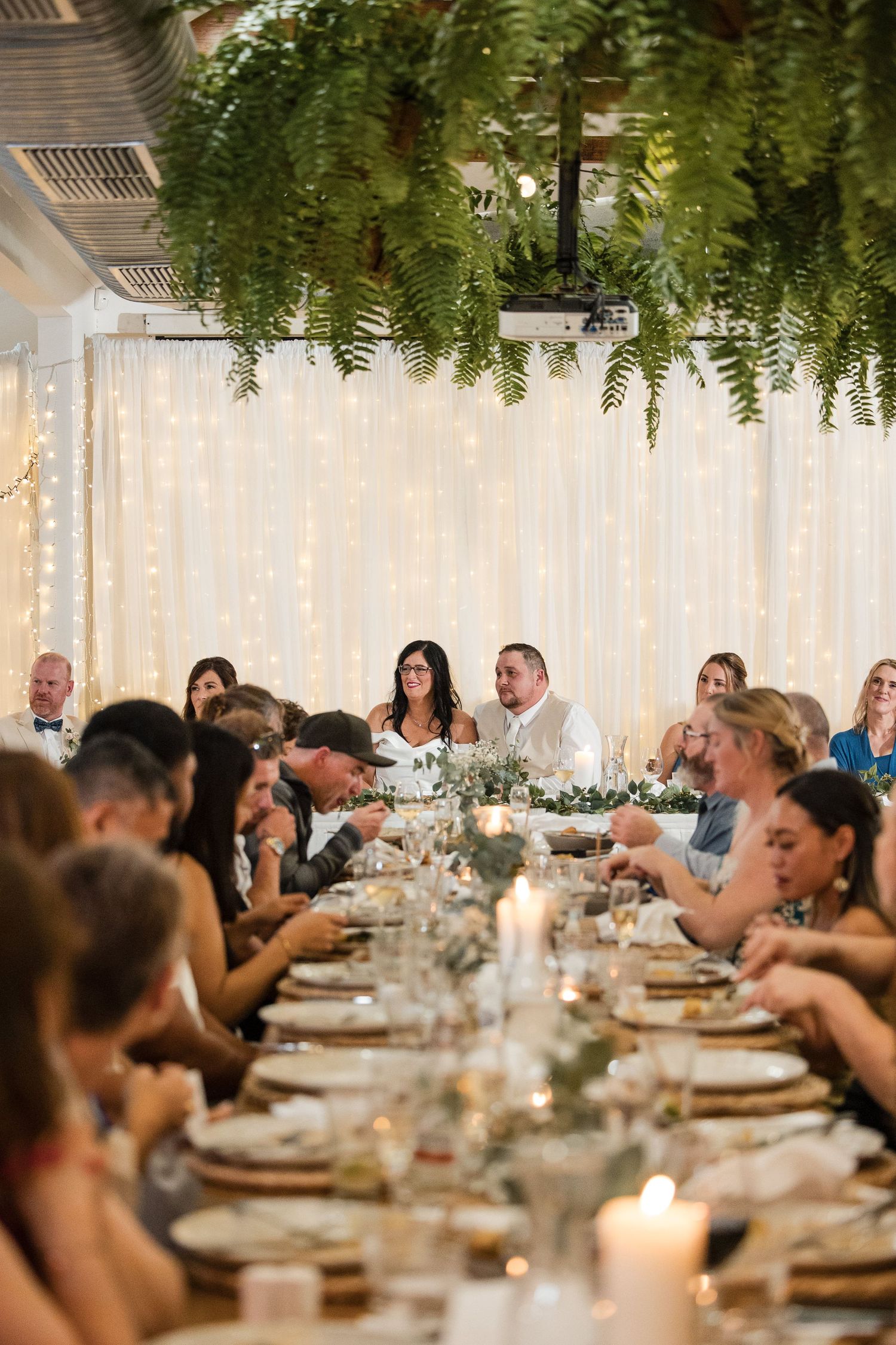 Elegant wedding reception with guests seated at a long decorated table, fairy lights backdrop, and lush greenery overhead.